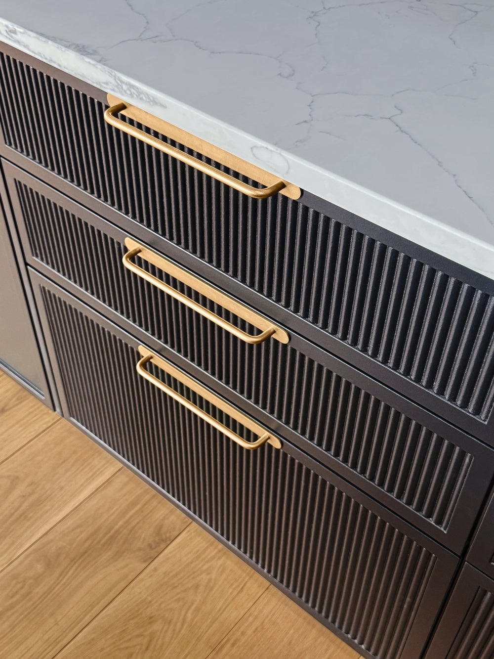 Shaker reeded kitchen cabinets with brass handles on a light wood floor.