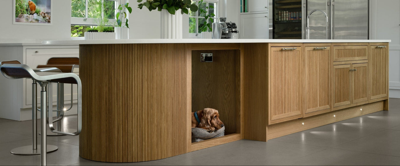 A kitchen island with a curved oak cabinet base and a white worktop, shown in a bright kitchen setting. A dog is visible inside the open lower cabinet area, and two bar stools sit beside the island.