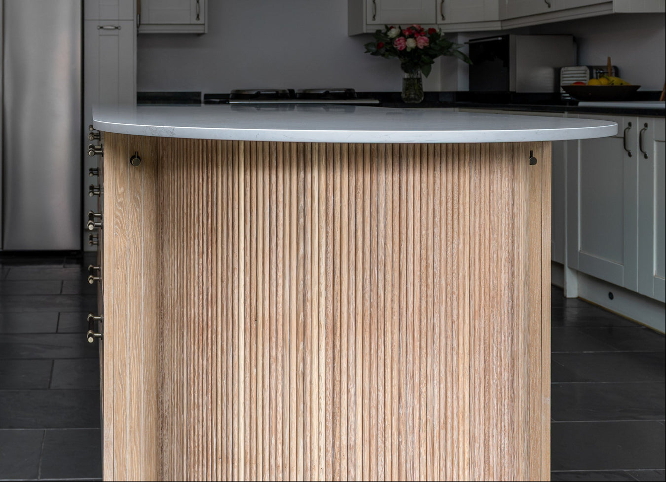 A modern kitchen island with a fluted light wood grain front and a curved white countertop, set in a contemporary kitchen with dark gray tile flooring and white cabinetry.