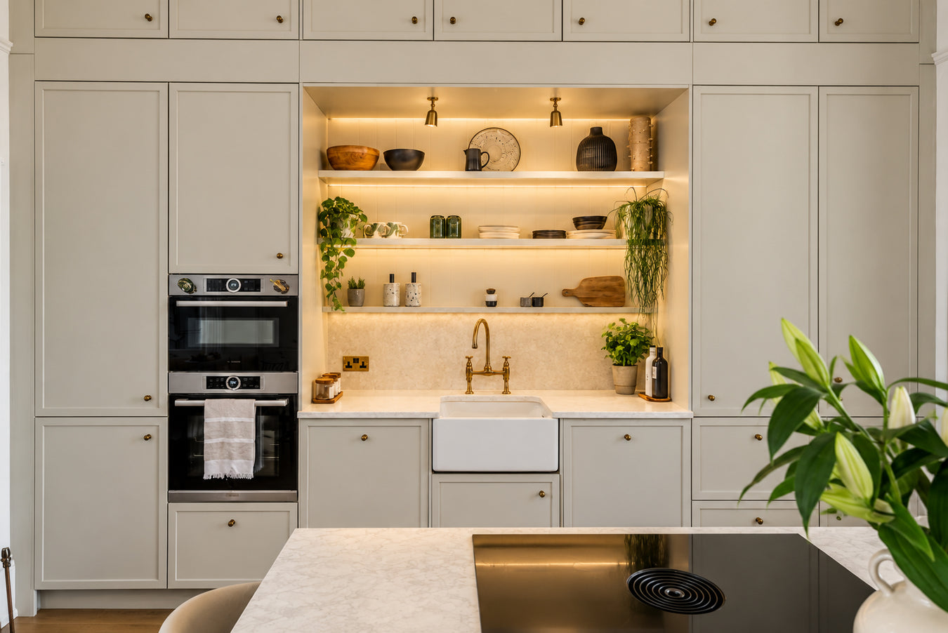 A modern kitchen interior with light beige cabinetry, built-in double ovens, a farmhouse sink, brass faucet, open shelving with decor and plants, and a dark cooktop in the foreground.