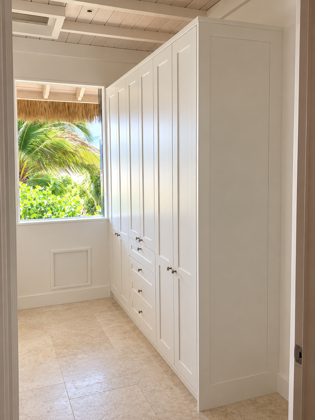 A long row of white shaker-style built-in wardrobe or closet doors with several drawers, shown in a bright room with tiled floor and a window looking out to palm trees.