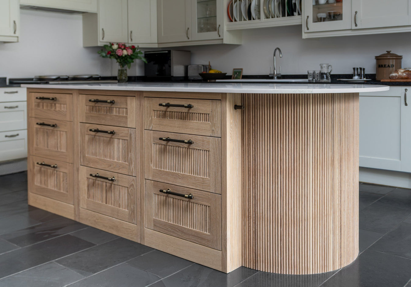A modern kitchen island with a curved fluted wood base, six drawers with dark metal handles, and a white countertop, set in a bright kitchen with gray floor tiles.