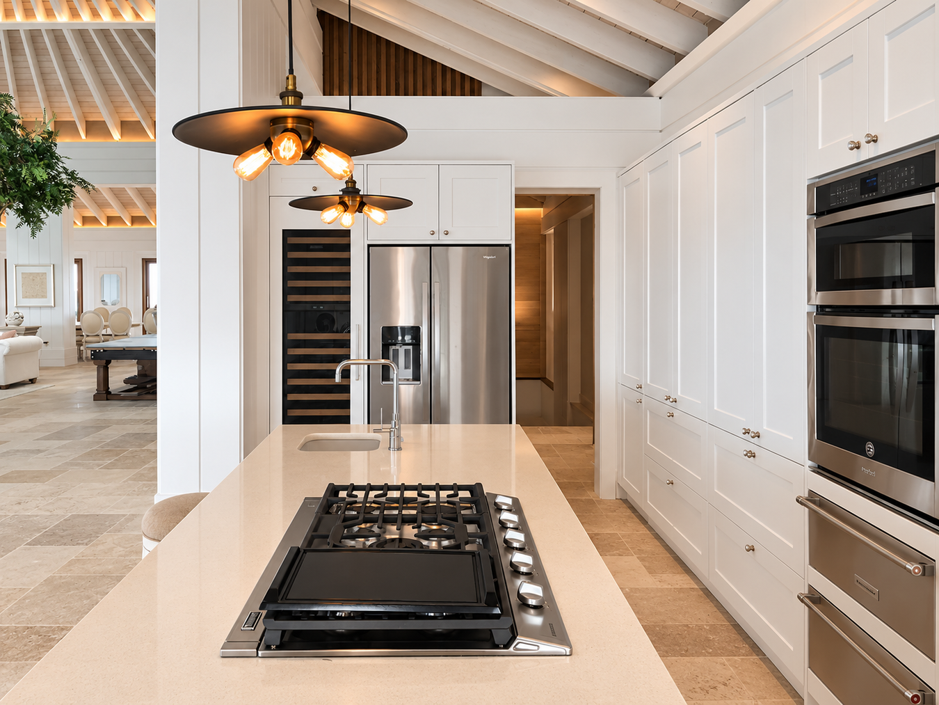 A modern shaker kitchen interior with white cabinetry, a cream island countertop, a black gas cooktop, stainless steel appliances including a double oven and refrigerator, and warm pendant lights hanging above the island.